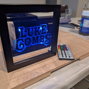 May include: Black framed LED sign with the text "LUKE COMBS" in blue neon. A remote control with multiple color options sits to the right of the sign. The sign is on a wooden surface.