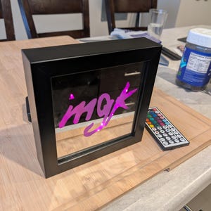 May include: A black framed LED light box with the word "mgk" in pink neon lettering. A remote control with multiple color options sits next to the light box on a wooden cutting board.