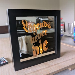 May include: A black framed mirror with the words "Legends never Die" in a gold-colored script. The mirror is illuminated from behind, creating a warm glow. The frame is rectangular and sits on a wooden surface.
