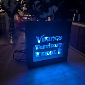 May include: A rectangular black light box featuring a glowing blue interior with the text Vikings Territory SKOL and a silhouette of the state of Minnesota. The device is powered by a black electrical cord extending from the side of the frame.