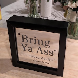 May include: A square black frame containing a clear glass pane with the text "Bring Ya Ass" printed in a bold, dark serif font, followed by the smaller text "-Anthony "Ant-Man" Edwards #5" in a cursive script style.