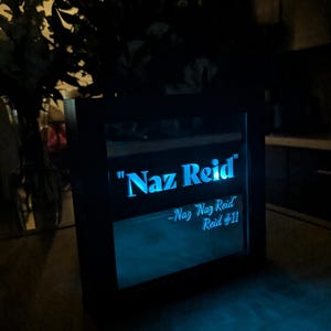 May include: A square black-framed light box featuring a glowing blue LED display. The text reads "Naz Reid" in a bold font, with the smaller script text "-Naz Naz Reid Reid #11" positioned directly below, emitting a bright cyan light.