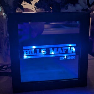 May include: A square black-framed LED light box featuring the text BILLS MAFIA in bold white capital letters, with the smaller text EST. 1960 positioned below. The display emits a bright blue glow, illuminating the lettering against a dark background.