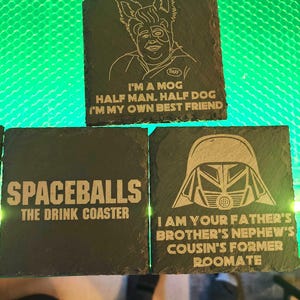 May include: Three black slate coasters with white engraved designs and text. One coaster features a MOG character with the text "I'M A MOG HALF MAN. HALF DOG I'M MY OWN BEST FRIEND". Another says "SPACEBALLS THE DRINK COASTER". The last one has a Darth Vader design and text.