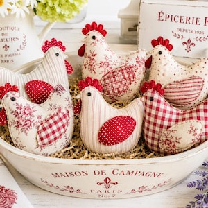 May include: A collection of six stuffed fabric chicken ornaments arranged in a large, shallow, cream-colored ceramic bowl labeled Maison de Campagne Paris No. 7. The chickens feature various red and white patterns, including floral, gingham, and polka dots.