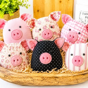 May include: Six plush pig ornaments in a wooden bowl. The pigs feature various fabric patterns including solid pink, floral, pink gingham, black with white polka dots, and pink striped fabric. Each pig has two small black button eyes and a pink felt snout.