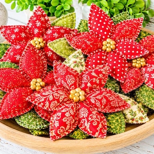 May include: A collection of fabric poinsettia ornaments in a wooden bowl. The flowers feature red and green patterned cotton petals with gold bead centres. The patterns include white snowflakes, polka dots, and floral motifs on red and green fabric.