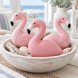 May include: Three plush flamingo figures in a white wooden bowl. Two flamingos feature a light pink floral pattern, while the centre flamingo has a light pink fabric with white polka dots. Each has a black beak tip and a small black eye.