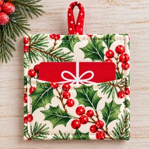 May include: A square fabric gift card holder featuring a festive holly and pine needle pattern on a cream background. The holder has a red and white polka dot fabric loop at the top and contains a red gift card with a white bow graphic.