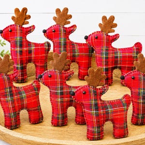 May include: A collection of six plush reindeer ornaments made from red, black, and yellow plaid fabric. Each reindeer features brown felt antlers, small black bead eyes, and a black pom-pom nose, with visible stitching along the edges of the fabric.