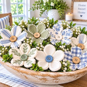May include: A large, weathered, light-colored wooden bowl filled with artificial greenery and several fabric flowers. The flowers feature various patterns including blue stripes, green solids, blue floral prints, and blue gingham, each centered with a button.