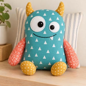 May include: A plush monster toy with a teal body featuring a white triangle pattern, two yellow horns with white dots, two orange arms with a white arrow pattern, and two yellow feet with white dots. It has two white eyes with black pupils and a smile.