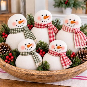 May include: Five plush snowman figures with orange carrot noses, black bead eyes, and rosy cheeks, each wearing a checkered scarf. The snowmen are nestled in a large wooden bowl filled with green pine branches, pinecones, and red berries.