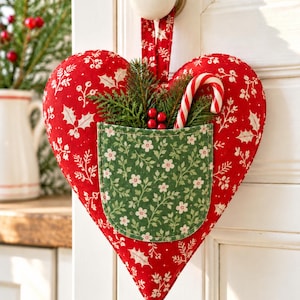 May include: A red heart-shaped fabric ornament with a white holly and berry print, featuring a green pocket with a white floral pattern. The pocket holds green pine sprigs, red berries, and a red and white striped candy cane. A fabric loop hangs from the top.
