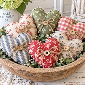 Può includere: Una collezione di sei cuori in tessuto imbottito in una ciotola di legno, ognuno con un motivo unico tra cui fiori rossi e bianchi, fiori verdi, quadretti vichy rossi e bianchi, righe blu e fiori blu, tutti decorati con un bottone in legno e spago.