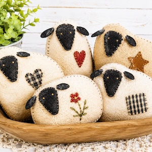 May include: Five stuffed sheep ornaments with cream-colored bodies and black felt faces. Each sheep features unique decorative accents including a red heart, a brown star, a black and white checkered heart, a red flower, and a black and white checkered patch.