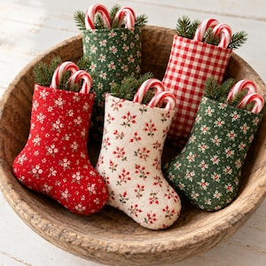 May include: Five small fabric stockings in red, green, and cream colors with floral and gingham patterns. Each stocking is filled with green pine sprigs and two red and white striped candy canes, arranged inside a large wooden bowl.