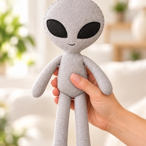 May include: A grey fabric alien doll featuring a large, rounded head with two prominent, smooth black oval eyes and a small, simple stitched mouth. The plush figure has a slender body, thin arms, and rounded feet, all crafted from a textured grey material.