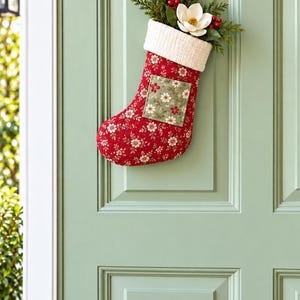 Peut inclure: Une chaussette de Noël en tissu rouge à motif floral blanc, avec un revers crème et un empiècement carré à motif floral vert. La chaussette est garnie de branches de pin vertes, de baies rouges et d'une fleur de magnolia blanche.