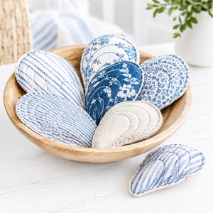 Puede incluir: Una colección de formas de conchas marinas cubiertas de tela en un cuenco de madera. Las conchas presentan varios patrones en azul y blanco, incluyendo rayas, estampados florales y diseños de puntos, con costuras visibles en los bordes.