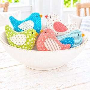 Puede incluir: Un conjunto de cinco adornos de pájaros de tela colocados en un cuenco blanco grande. Los pájaros presentan varios patrones que incluyen lunares, estampados florales y rayas en tonos azul, verde, rosa y blanco, cada uno con un pequeño pico naranja.