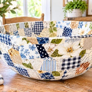 May include: A large, round decorative bowl featuring a patchwork design of various fabric patterns, including blue and white gingham, floral prints, polka dots, and striped textures in shades of navy, light blue, white, and tan.