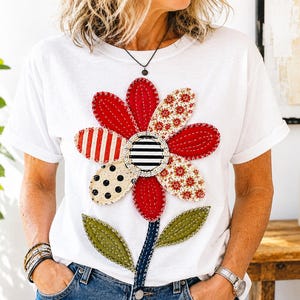 Könnte beinhalten: Ein weißes Kurzarm-T-Shirt mit einer großen, zentralen Blumenapplikation aus verschiedenen Stoffstücken. Die Blütenblätter zeigen rote, gepunktete, gestreifte und florale Muster mit sichtbaren weißen Nähten. Ein dunkelblauer Stiel verläuft nach unten.
