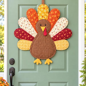 May include: A fabric turkey wall hanging featuring a brown body, yellow beak, red wattle, and black eyes. The tail feathers are made of quilted fabric patches in orange, yellow, and white floral patterns, along with a yellow and white gingham check.