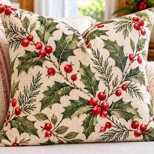 May include: A square throw pillow featuring a cream-colored fabric base with a repeating botanical print of green holly leaves, pine needles, and bright red berries. The design covers the entire front surface of the pillow in a dense pattern.