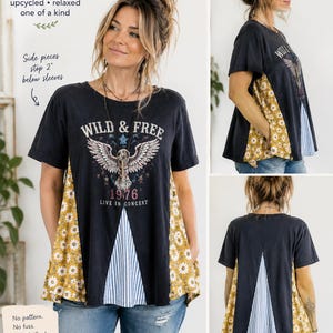May include: A black short-sleeve swing tee featuring a graphic of an eagle with the text WILD & FREE 1976 LIVE IN CONCERT. The shirt has side panels with a yellow daisy print and a central blue and white striped panel, with side pieces stopping 2 inches below the sleeves.