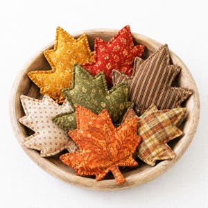 May include: A collection of stuffed fabric maple leaves in various autumn colors and patterns, including mustard yellow, deep red, olive green, brown stripes, cream with small dots, orange floral, and plaid, all arranged inside a round wooden bowl.