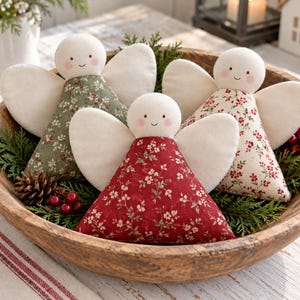 May include: Three fabric angel ornaments with round white heads, pink cheeks, and simple stitched faces. Each angel has cream-colored fabric wings and a triangular body featuring floral patterns in shades of olive green, deep red, and cream.