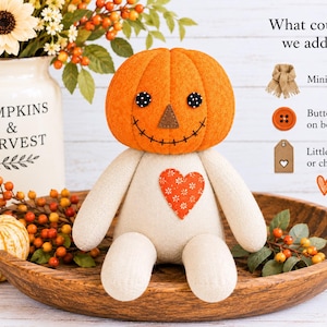 May include: A fabric doll with an orange pumpkin head, black button eyes, a brown triangular nose, and a stitched smile. The body is off-white with an orange heart patch featuring a white floral pattern. Text reads: What could we add?