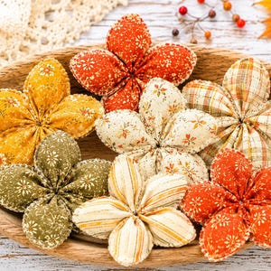 May include: A collection of six fabric flowers in a wooden bowl. The flowers feature various patterns including floral prints, plaid, and stripes in autumn colors such as orange, yellow, olive green, and cream. Each flower has five petals.