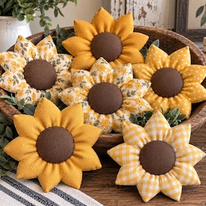 Puede incluir: Un conjunto de seis adornos de tela con forma de girasol, con centros circulares de color marrón oscuro y pétalos en varios patrones amarillos, incluyendo lisos, lunares blancos, cuadros vichy amarillos y estampados florales, en un cuenco de madera.