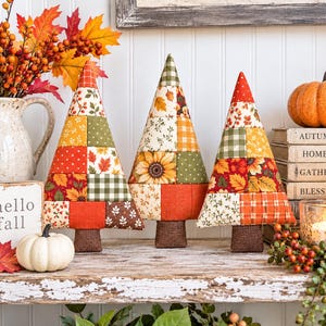 Puede incluir: Tres árboles de tela en forma de cono con un diseño de patchwork de patrones otoñales que incluyen girasoles, hojas y cuadros en tonos naranja, amarillo, verde y marrón, cada uno con una pequeña base rectangular marrón.