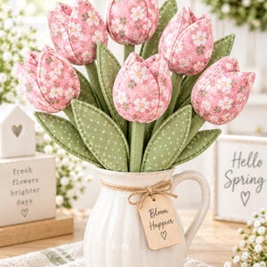 Könnte beinhalten: Ein Strauß aus Stofftulpen in Rosatönen mit weißem Blumenmuster und grünen Blättern mit Punkten. Die Tulpen sind in einem weißen Keramikkrug mit vertikalen Rillen arrangiert, der mit einer Schnur und einem Holzanhänger mit Aufschrift versehen ist.