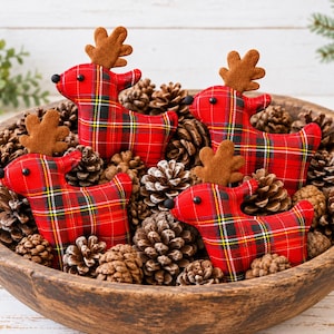 May include: Four plush reindeer ornaments featuring a red, black, white, and yellow tartan pattern. Each reindeer has brown felt antlers and a small black bead for an eye. The reindeer are nestled inside a large, round, dark brown wooden bowl filled with numerous natural brown pinecones.