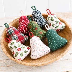 May include: A collection of eight fabric bell ornaments featuring various patterns including red and green plaid, white with holly berries, dark green with white dots, and red with white floral designs, each with a small fabric hanging loop.