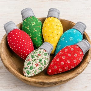 May include: A collection of six fabric ornaments shaped like vintage light bulbs, each featuring a unique pattern including polka dots, holly leaves, and snowflakes in red, green, yellow, and blue, all resting inside a round wooden bowl.