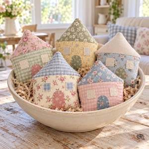 Puede incluir: Un conjunto de cinco adornos de tela acolchada con forma de casa dentro de un cuenco de cerámica. Cada casa presenta una combinación única de textiles con estampados florales, de cuadros y lisos, con ventanas y puertas arqueadas cosidas.