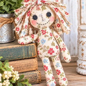 Puede incluir: Una muñeca de tela con una base de color crema con un estampado floral de flores rojas y azules con hojas verdes. La muñeca tiene ojos de botón negros, nariz y boca cosidas y mejillas rosadas. El cabello está hecho de tiras de tela deshilachadas.