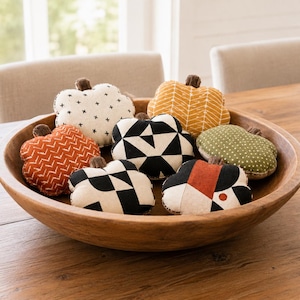 Op de afbeelding: Een grote, ronde houten kom gevuld met zeven kleine, opgevulde stoffen pompoenen. De pompoenen hebben verschillende patronen, waaronder zwart-witte geometrische vormen, oranje-wit chevron, mosterdgeel visgraatmotief en groene stippen.