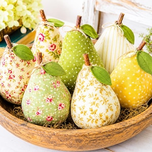 May include: A collection of seven fabric pears in a round wooden bowl. Each pear features a unique pattern, including floral prints, polka dots, and stripes in shades of green, yellow, and cream. Each pear has a brown twig stem and a green leaf.