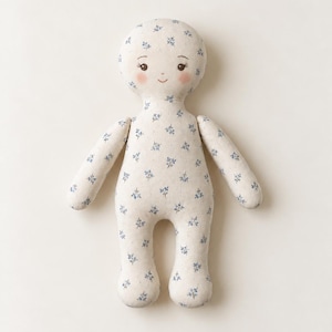 May include: A soft fabric doll featuring a cream-coloured base with a delicate blue floral pattern. The doll has a rounded head, simple arms and legs, and a printed face with dark eyes, arched eyebrows, a small smile, and soft pink rosy cheeks.