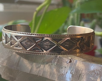 May include: A silver bracelet with a geometric pattern of black diamonds and arrows. The bracelet is made of metal and has a polished finish.