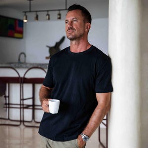May include: A black crew neck t-shirt. The person is holding a white mug. The person is wearing a silver watch. The person is leaning against a white column. The background includes a kitchen area with a bar and artwork.