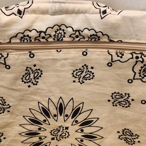 May include: A cream-colored fabric bag featuring a black paisley bandana print pattern. The bag includes a horizontal zipper closure across the top with a gold-toned circular metal ring pull attached to the zipper slider for easy opening and closing.