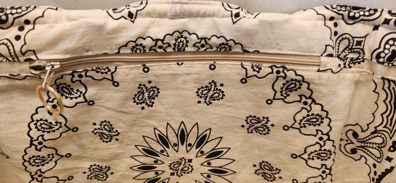 May include: A cream-colored fabric bag featuring a black paisley bandana print pattern. The bag includes a horizontal zipper closure across the top with a gold-toned circular metal ring pull attached to the zipper slider for easy opening and closing.