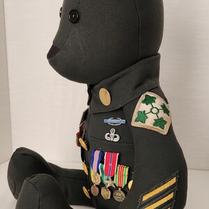 Op de afbeelding: Een donkergroene teddybeer met een militair uniform. De beer heeft een zwart knoopoog en een bruine neus. Het uniform bevat patches, medailles en strepen, wat duidt op een gedecoreerd lid. De beer zit.